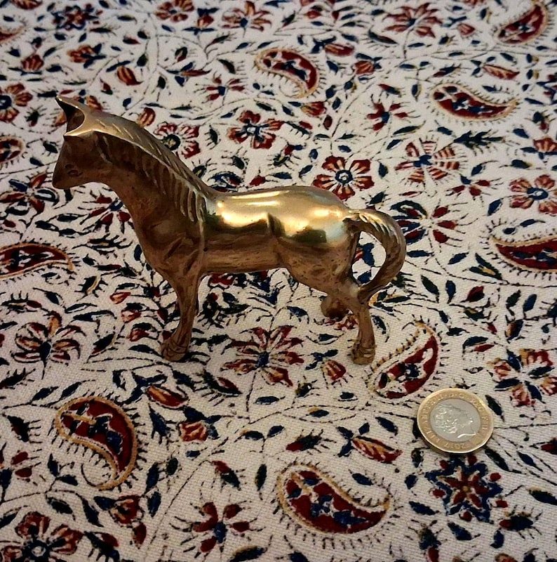May include: A small, brass horse figurine in a standing pose. The horse has a detailed mane and tail, and is set against a patterned fabric with floral and paisley designs. A coin is next to the horse.