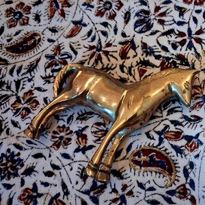 May include: A brass horse figurine in a trotting pose. The sculpture is highly polished and reflects light, with details in the mane, tail, and legs. The horse is set against a patterned fabric background with floral and paisley designs.