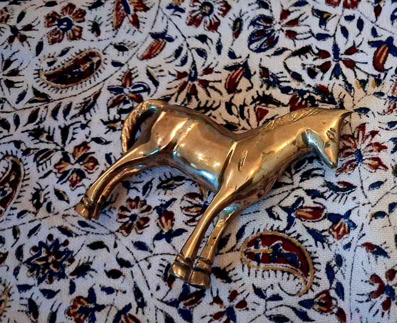 May include: A brass horse figurine in a trotting pose. The sculpture is highly polished and reflects light, with details in the mane, tail, and legs. The horse is set against a patterned fabric background with floral and paisley designs.