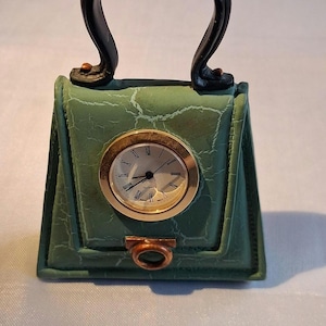 May include: A green handbag-shaped desk clock with a cracked texture finish. The clock face is round with a gold-toned bezel, black hands, and Roman numerals. It features a black arched handle and a small copper-coloured ring detail at the base.