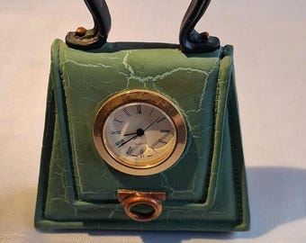 May include: A green handbag-shaped desk clock with a cracked texture finish. The clock face is round with a gold-toned bezel, black hands, and Roman numerals. It features a black arched handle and a small copper-colored ring detail at the base.