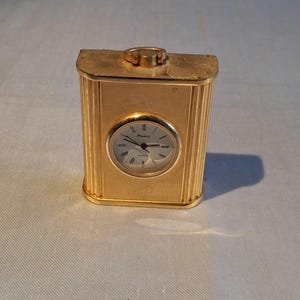 May include: A gold-toned rectangular quartz travel clock with a metallic finish and vertical ribbed side panels. The clock face features black Roman numerals, thin black hands, and the brand name Premia printed in the center of the white dial.