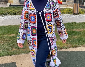 May include: A long, open-front crochet cardigan featuring colorful square patterns. The white trim and fringe add detail to the garment. A white tie closure and tassels hang from the front. The cardigan is worn with dark blue jeans and white sneakers.