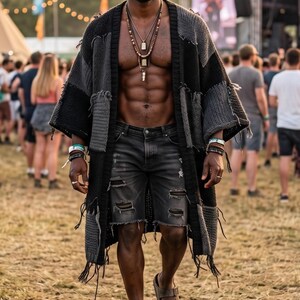 May include: A long, open-front patchwork cardigan featuring alternating black and charcoal gray knit panels with frayed edges and fringe trim. The garment is worn over distressed black denim shorts and brown double-strap slide sandals.