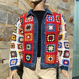 May include: Denim jacket with a red front panel and crochet sleeves. The crochet features colorful squares in various hues, including blue, yellow, and green. The jacket has a classic collar and button closure, displayed on a mannequin.