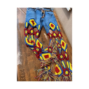 May include: Light blue denim jeans with colorful crochet detailing. The crochet design features red, yellow, and blue geometric shapes and fringe, creating a unique and artistic look. A brown belt is threaded through the belt loops.