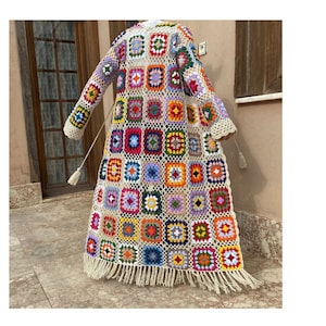 May include: A long, open-front crochet cardigan featuring a colorful granny square pattern in various hues including red, blue, yellow, and purple. The garment has long sleeves, a cream-colored lattice border, and a fringed hemline.