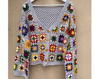 May include: A gray, crocheted cardigan with a V-neck and long sleeves. The cardigan features colorful, square granny squares in various colors, including red, yellow, blue, and green. It has wooden buttons down the front and is hanging on a white hanger.