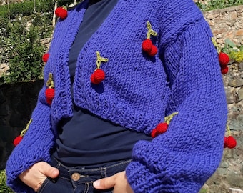 May include: A cropped, royal blue knit cardigan adorned with red cherry appliqués. The cardigan features long sleeves and a V-neck design. The cherries have green stems and are evenly spaced across the garment.
