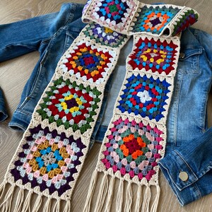 May include: A colorful, handmade crochet scarf featuring square granny stitch patterns in various colors. The scarf has a cream-colored border and fringe. The scarf is draped over a denim jacket.