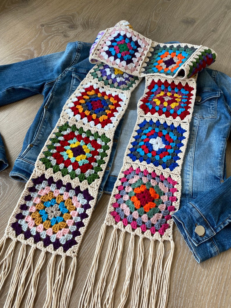 May include: A colorful, handmade crochet scarf featuring square granny stitch patterns in various colors. The scarf has a cream-colored border and fringe. The scarf is draped over a denim jacket.