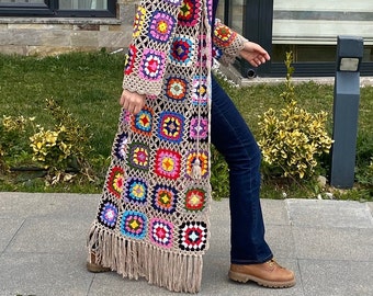 Könnte beinhalten: Lange, offene Häkeljacke in Beige mit bunten quadratischen Mustern. Die Strickjacke hat lange Ärmel, einen Fransenbesatz und ein Quastendetail. Das Kleidungsstück wird über einem dunklen Oberteil und einer blauen Jeans getragen.