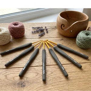 Peut inclure: Ensemble de crochets avec des manches gris et des pointes dorées, disposés en éventail. L'image comprend également des pelotes de fil roses et vertes, un bol à fil en bois et des brins de lavande, le tout sur une surface en bois.