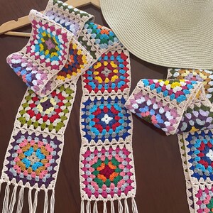 May include: A colorful, handmade crochet scarf with a granny square design. The scarf features squares in various colors, including pink, blue, green, and yellow, with a beige border and fringe. A straw hat rests nearby on a wooden hanger.