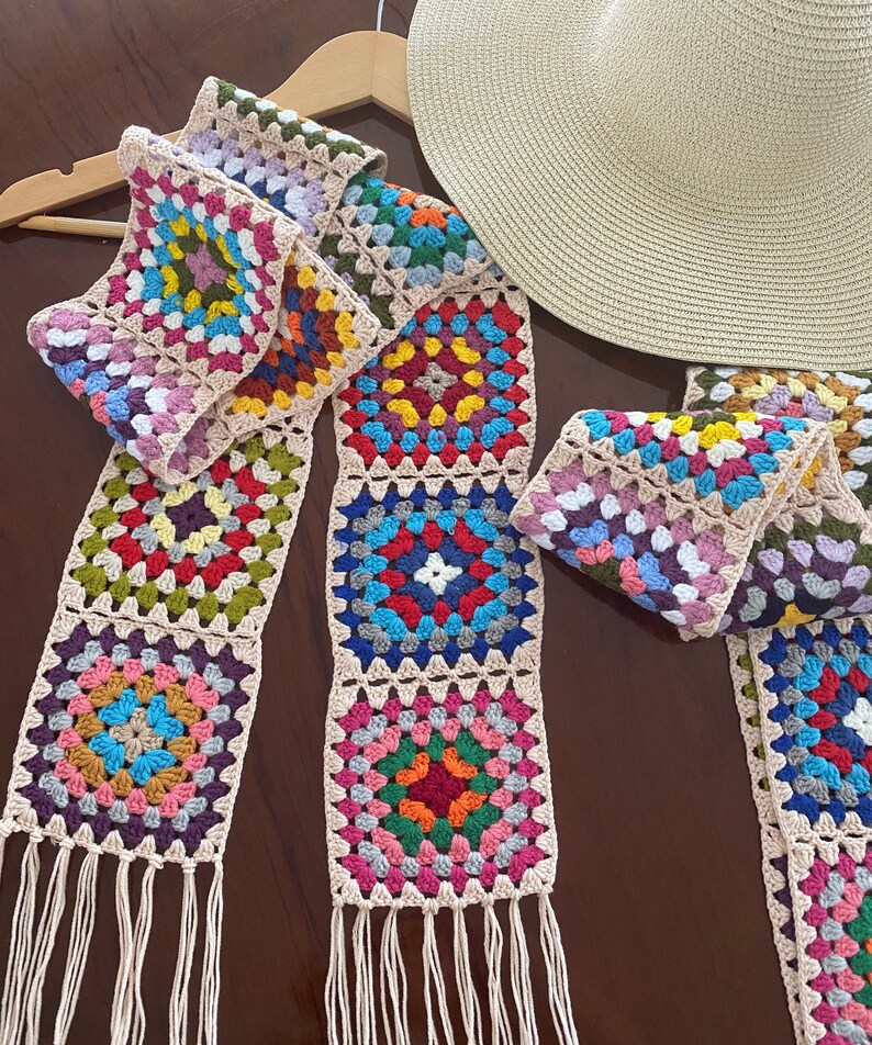 May include: A colorful, handmade crochet scarf with a granny square design. The scarf features squares in various colors, including pink, blue, green, and yellow, with a beige border and fringe. A straw hat rests nearby on a wooden hanger.