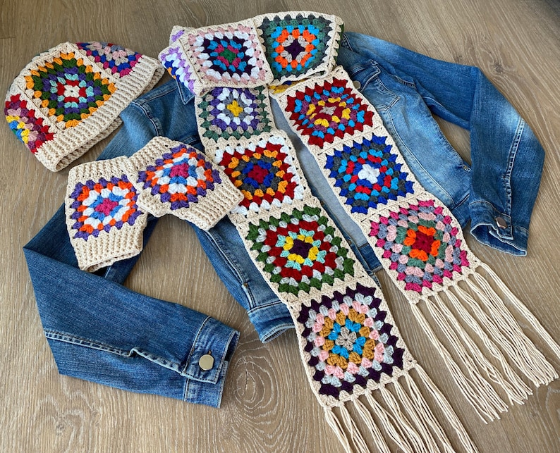 May include: A crochet set featuring a hat, fingerless gloves, and a scarf. The items are made with beige yarn and colorful square patterns. The scarf has fringe at the ends. The set is displayed on a denim jacket.