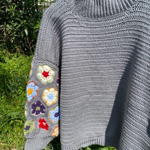 May include: A gray crochet sweater with a high neck and short sleeves. The left sleeve features a colorful floral pattern in red, blue, yellow, and purple. The sweater is hanging on a white hanger against a backdrop of green foliage.