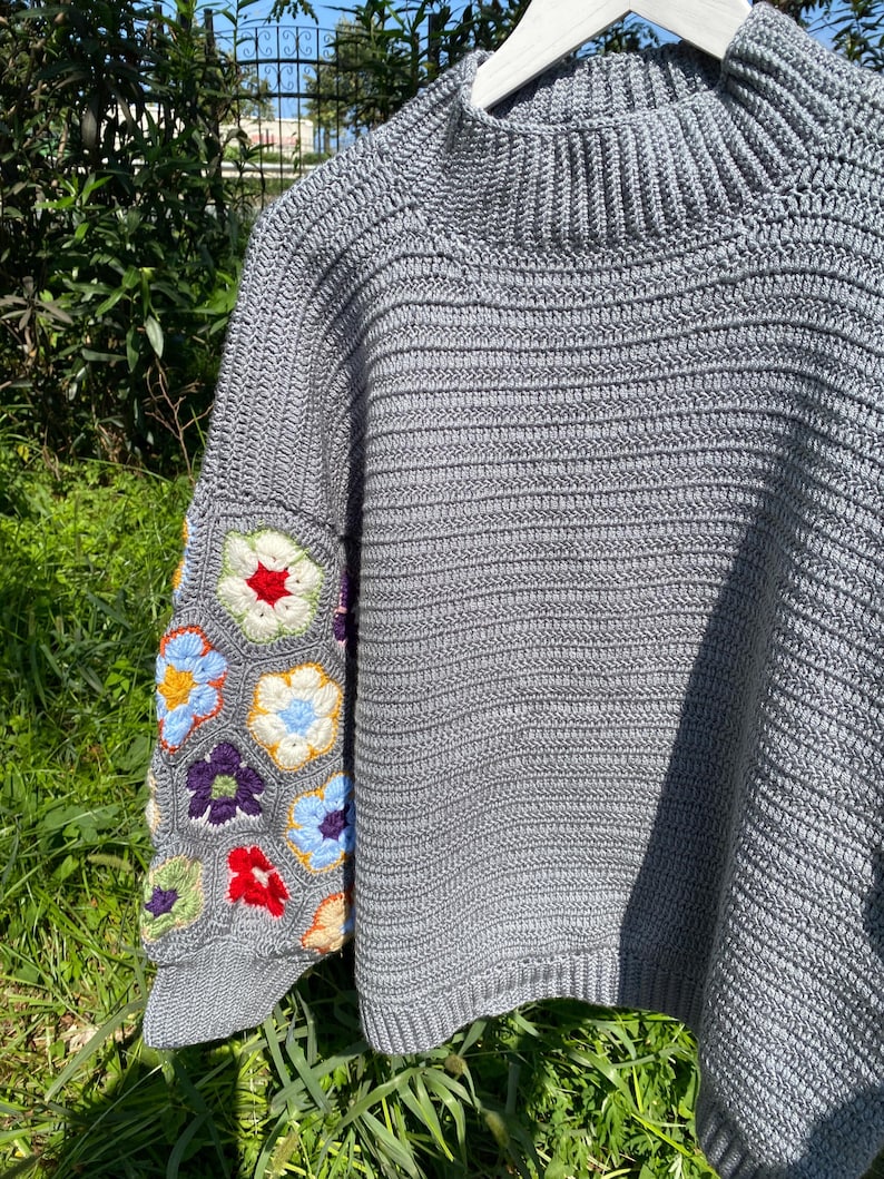 May include: A gray crochet sweater with a high neck and short sleeves. The left sleeve features a colorful floral pattern in red, blue, yellow, and purple. The sweater is hanging on a white hanger against a backdrop of green foliage.