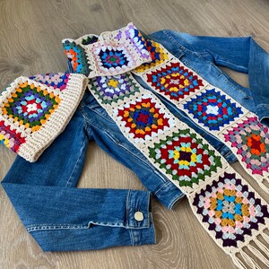May include: A crocheted hat and scarf set with a colorful granny square design. The hat and scarf feature a cream-colored border and fringe, with squares in various colors like red, blue, green, and purple. The set is displayed on a denim jacket.