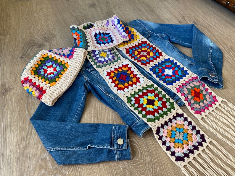 May include: A crocheted hat and scarf set with a colorful granny square design. The hat and scarf feature a cream-colored border and fringe, with squares in various colors like red, blue, green, and purple. The set is displayed on a denim jacket.