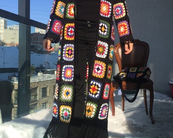 May include: A long, black crochet cardigan with colorful square patterns. The cardigan features a fringe hem and is worn with black pants and brown boots. A vintage chair and a matching bag are in the background.