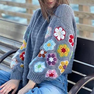 May include: A gray crochet sweater with colorful floral patterns on the sleeves. The flowers are in shades of red, white, yellow, blue, and purple. The sweater has a ribbed hem and cuffs.