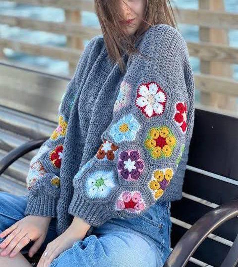May include: A gray crochet sweater with colorful floral patterns on the sleeves. The flowers are in shades of red, white, yellow, blue, and purple. The sweater has a ribbed hem and cuffs.