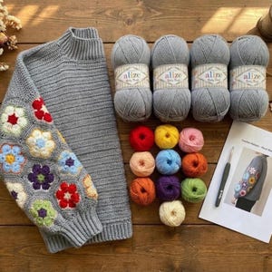 May include: A gray sweater with a floral crochet sleeve, accompanied by gray yarn balls, colorful yarn skeins, a crochet hook, and a pattern sheet. The sweater features a high neck and ribbed cuffs.