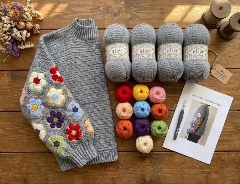 May include: A gray sweater with a floral crochet sleeve, accompanied by gray yarn balls, colorful yarn skeins, a crochet hook, and a pattern sheet. The sweater features a high neck and ribbed cuffs.