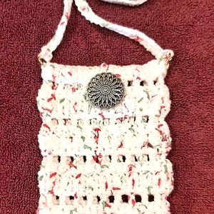 May include: A white crocheted bag with a red and green floral pattern. The bag has a long strap and a silver button closure.