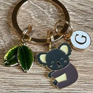 May include: Gold key ring with a koala charm, two green leaves, and a charm with the letter "G" on a white background.