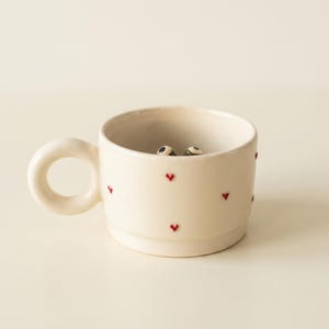 Può includere: Una tazza in ceramica color crema con un manico rotondo e piccoli cuori rossi sparsi sull'esterno. All'interno della tazza sono visibili due piccoli occhi dall'aspetto realistico che spuntano dal fondo del recipiente.