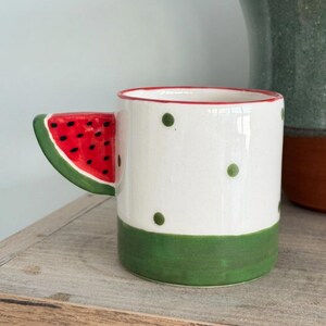 Può includere: Una tazza in ceramica bianca con base verde e piccoli pois verdi. Il manico ha la forma di una fetta di anguria, dipinta con un interno rosso, semi neri e un bordo verde, creando un design distintivo a tema frutta.