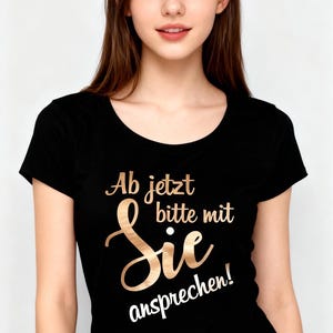 Op de afbeelding: Zwart T-shirt met ronde hals en gouden tekst "Ab jetzt bitte mit Sie ansprechen!". Het T-shirt wordt gedragen door een persoon met lang bruin haar.
