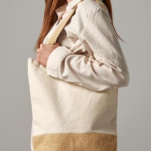Könnte beinhalten: Eine weiße Canvas-Tragetasche mit braunem Jute-Boden. Die Tasche hat einen langen Riemen und wird von einer Person getragen.