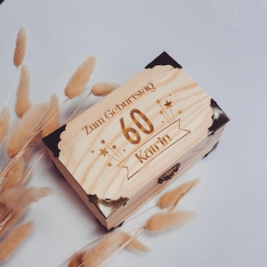 May include: A light-coloured wooden box with dark corner accents. The box is engraved with the words "Zum Geburtstag", the number "60", and the name "Katrin". Decorative stars and a banner are also engraved on the box.