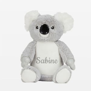 Könnte beinhalten: Ein kuscheliges graues Koala-Bär-Spielzeug mit weißen Akzenten an Ohren und Füßen. Der Koala hat eine schwarze Nase und kleine schwarze Augen. Der Name "Sabine" ist in grauer Schrift auf dem weißen Bauch des Stofftiers gestickt.