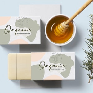 Puede incluir: Dos cajas blancas con un dise&ntilde;o abstracto verde y rosa y el texto "Organic Handmade Soap". Las cajas est&aacute;n sobre una superficie azul claro con una peque&ntilde;a rama de hojas verdes y un cuenco de miel con un cuchar&oacute;n de miel.