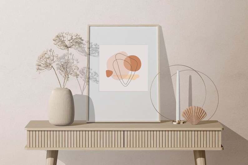 Puede incluir: Una impresi&oacute;n art&iacute;stica abstracta enmarcada con formas y l&iacute;neas en tonos tierra se encuentra sobre una consola de madera clara. Un jarr&oacute;n beige con flores secas, una vela y una concha decorativa completan la decoraci&oacute;n minimalista.