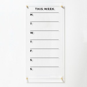A weekly planner with black text on a clear acrylic and 4 gold standoffs around the board. Each day has its own blank are to write in the notes, menu, to-dos, goals etc. Behind the planner is a white wall which looks elegant when combined.