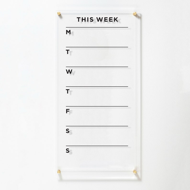 A weekly planner with black text on a clear acrylic and 4 gold standoffs around the board. Each day has its own blank are to write in the notes, menu, to-dos, goals etc. Behind the planner is a white wall which looks elegant when combined.