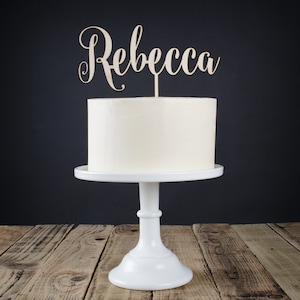 Può includere: Una torta bianca glassata su un'alzatina bianca con un cake topper in legno "Rebecca". La torta è posta su uno sfondo scuro. L'alzatina è su una superficie di legno.