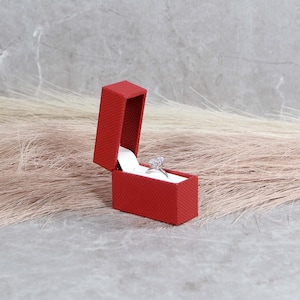 May include: A red ring box with a white interior, open and displaying a silver ring with a clear stone.