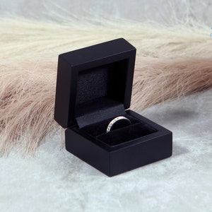 May include: A black ring box with a velvet interior holds a diamond ring. The ring is set with multiple small diamonds. A pink rose and other flowers are in the background.