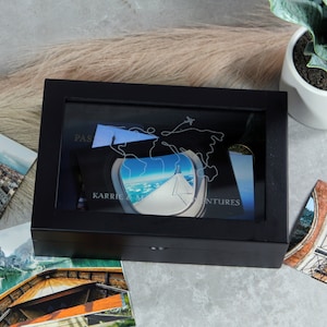 May include: Black wooden box with a glass top. The box has a world map design with the text "PASSPORT TO ADVENTURES" and a picture of an airplane window view. The box is surrounded by postcards.