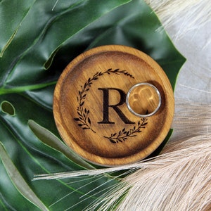 May include: A wooden ring dish with a carved wreath and the letter "R" in the center. A silver ring is resting on the dish. The dish is on a green leaf and a beige background.