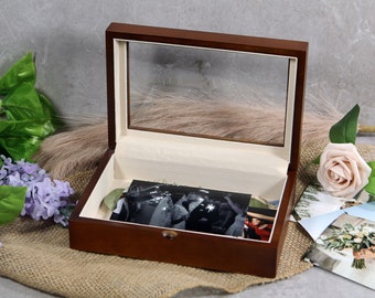 May include: A wooden keepsake box with a clear glass top, open to reveal photographs. The box is brown with a cream-colored interior. A few photos are visible inside the box and next to it, along with a pink rose and purple flowers.
