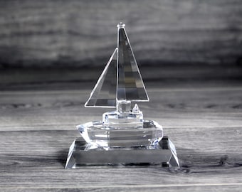 Puede incluir: Una figurita de velero de cristal transparente con una vela y un casco detallados. El velero está sobre una base de cristal transparente.