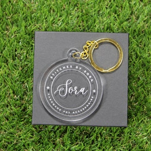 Puede incluir: Un llavero de acr&iacute;lico transparente con un anillo de metal dorado. El llavero tiene un c&iacute;rculo blanco con el texto "Stitches by Sora" y el nombre "Sora" escrito en cursiva.