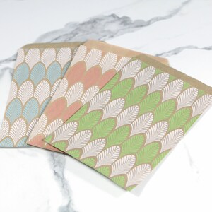 K&ouml;nnte beinhalten: Drei Papier-Geschenkbeutel mit braunem Hintergrund und einem wei&szlig;en und gr&uuml;nen geometrischen Muster. Die Beutel sind &uuml;bereinander gestapelt, wobei der gr&uuml;ne Beutel oben, der rosa Beutel in der Mitte und der blaue Beutel unten liegt.